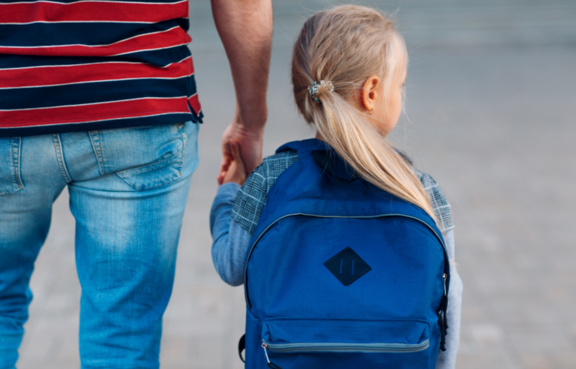 Un parent tient la main de son enfant qui porte un cartable, sur le chemin de l'école